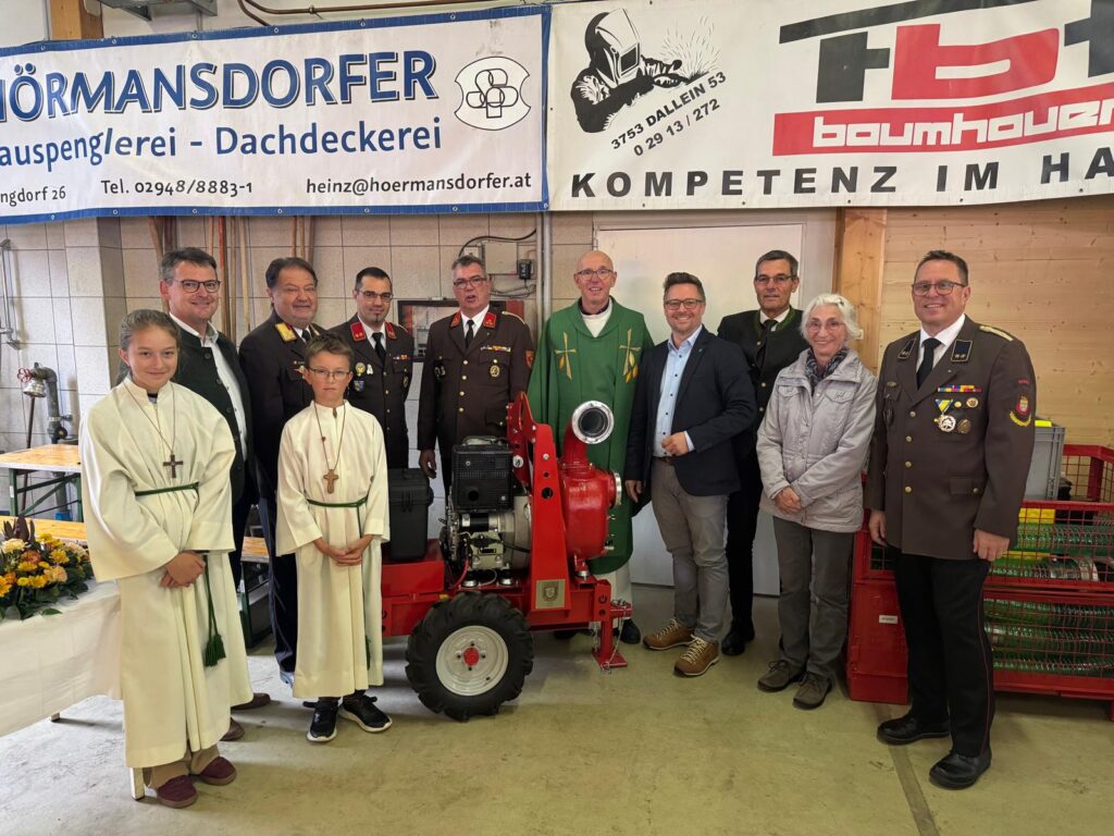 Vertreter der Freiwilligen Feuerwehr Weitersfeld, Feuerwehrfunktionäre, Ehrengäste und zwei Ministranten stehen nach der Segnung der neuen Pumpe SPA 150 gemeinsam für ein Gruppenfoto im Feuerwehrhaus.