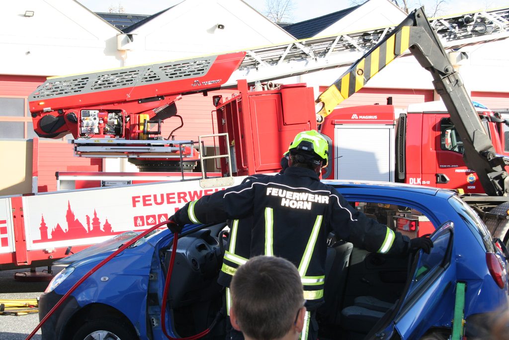 Ein Feuerwehrmitglied demonstriert an einem blauen Pkw den Einsatz eines hydraulischen Rettungsgeräts,