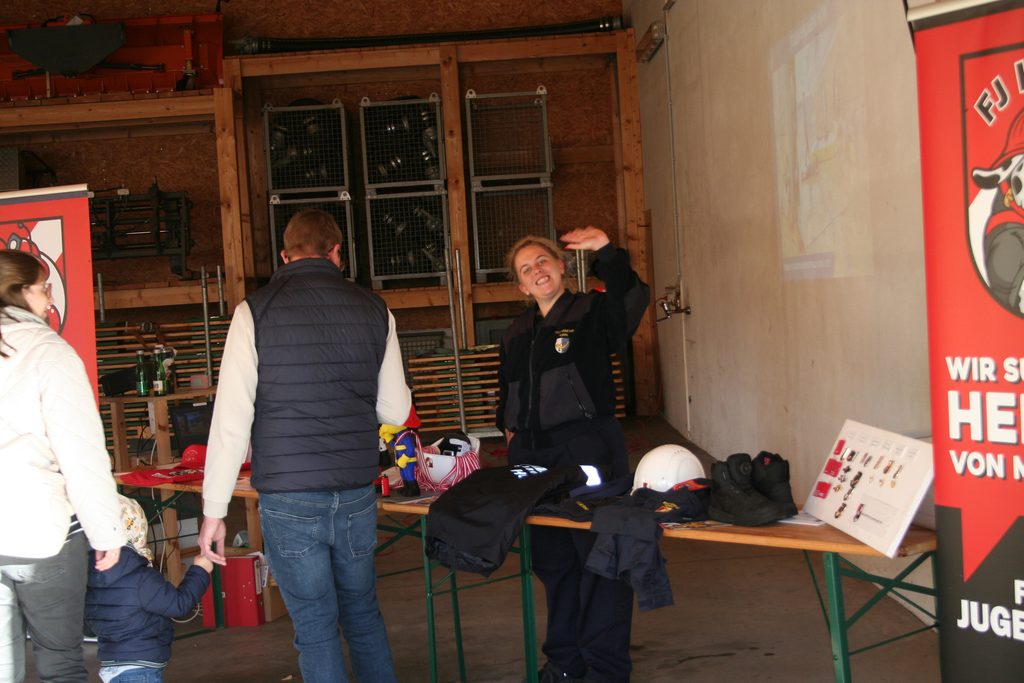 Ein Feuerwehrmitglied informiert Besucher an einem Tisch über Ausrüstungsgegenstände und Bekleidung der Feuerwehr.